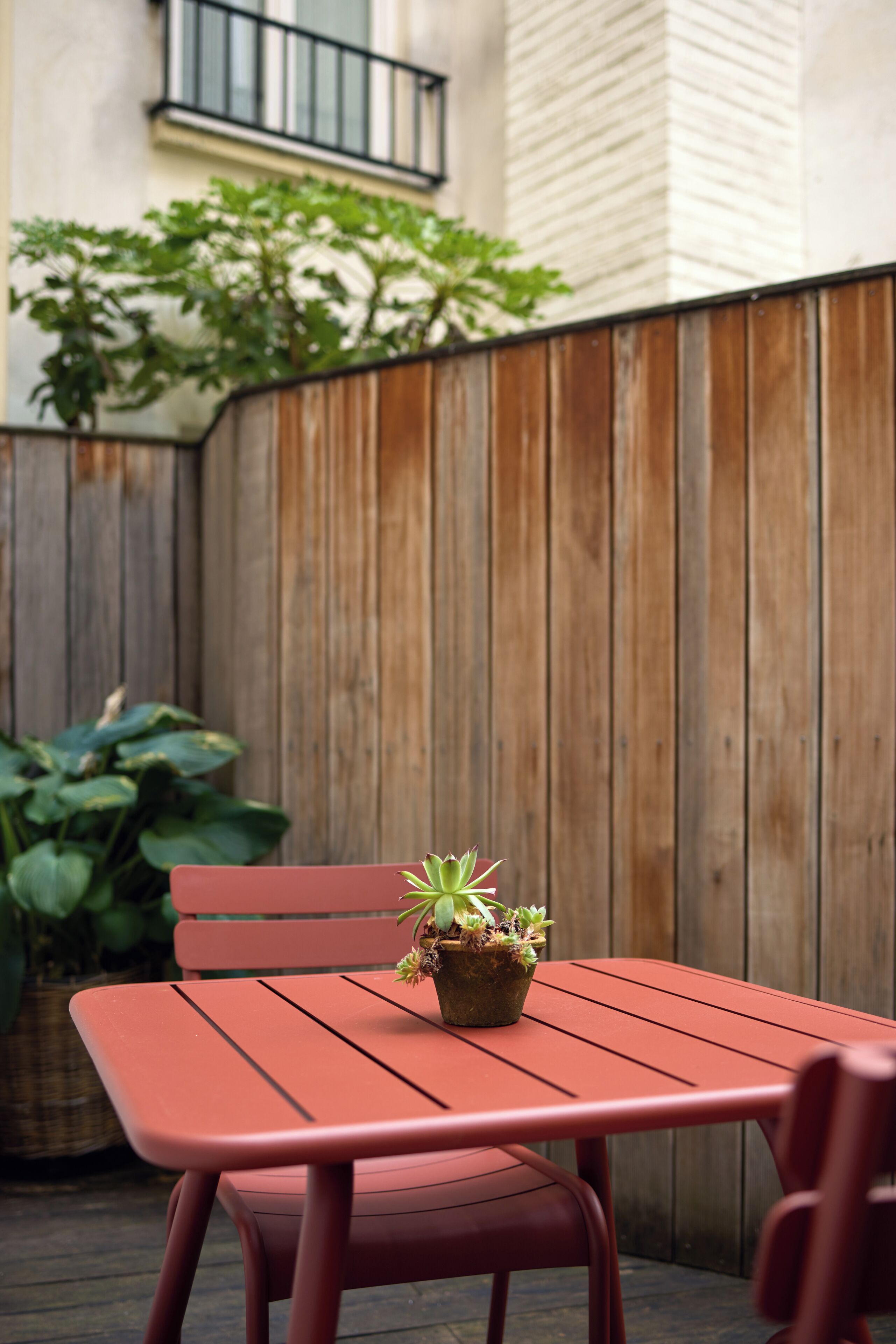 A red metal table and chair set in an outdoor area with a wooden fence and a potted plant.