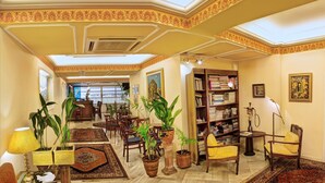 A spacious living room with a high ceiling, ornate ceiling design, multiple potted plants, a bookshelf, and a large window providing natural light. - Nicola Hotel (Athens)