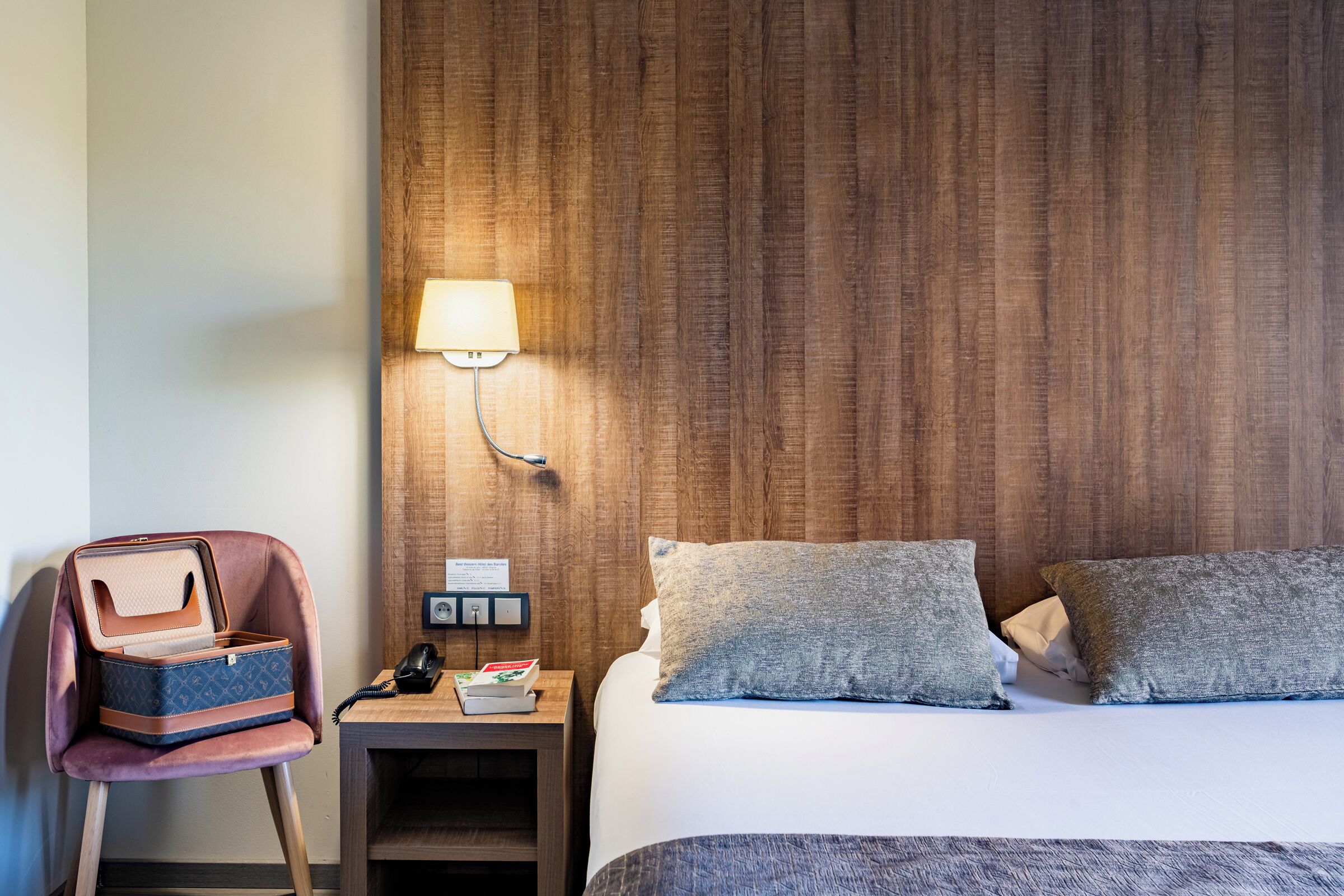 A hotel room with a bed, a nightstand with a lamp, a chair with a suitcase, and a wooden headboard.