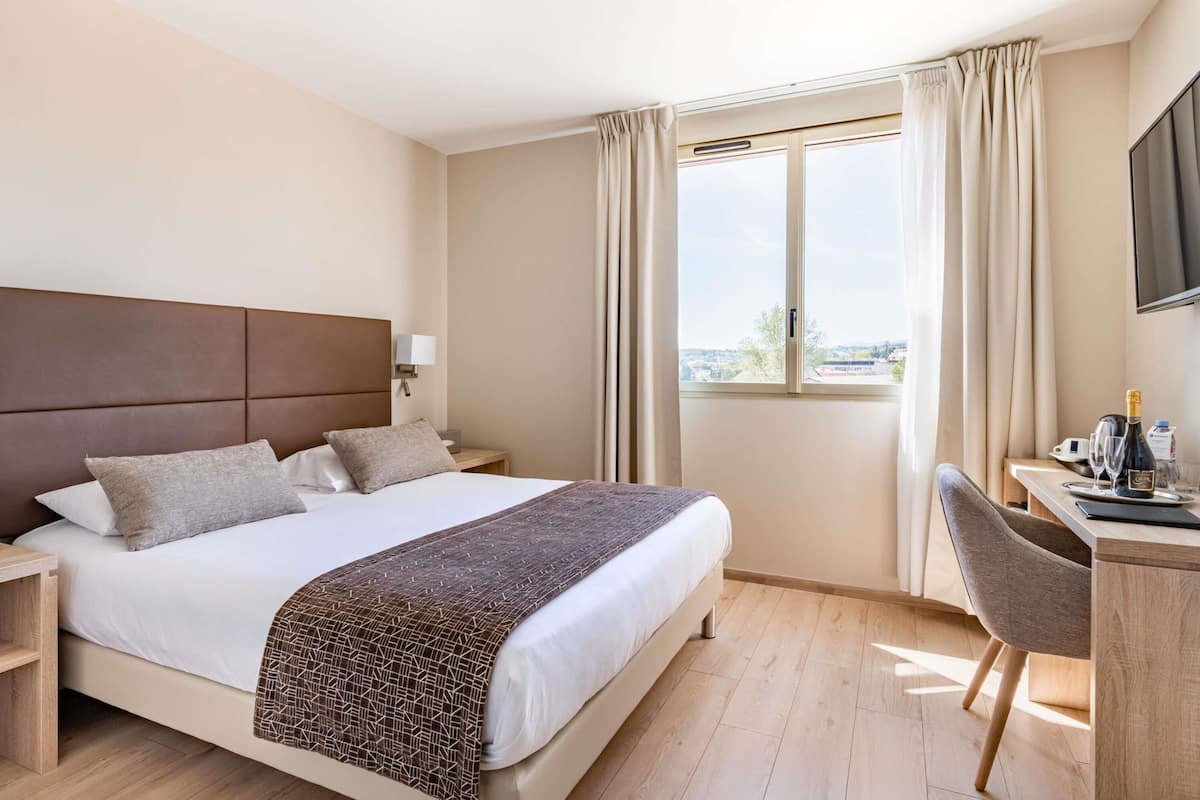 Une chambre d’hôtel avec un grand lit, un bureau avec une chaise, une télévision et une fenêtre avec des rideaux.