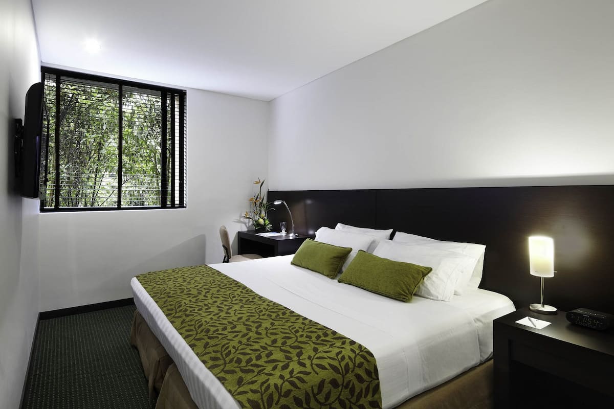 Une chambre d’hôtel avec un lit, une table de chevet, une lampe et une fenêtre donnant sur des arbres.