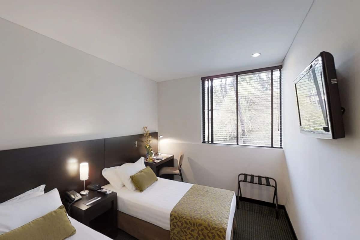 Une chambre d’hôtel avec deux lits, un bureau, une chaise, une télévision et une fenêtre avec des stores.