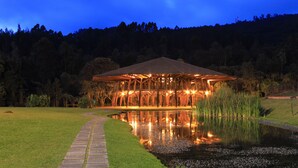Um pavilhão com telhado sustentado por vigas de madeira, cercado por gramado e um lago que reflete a estrutura.