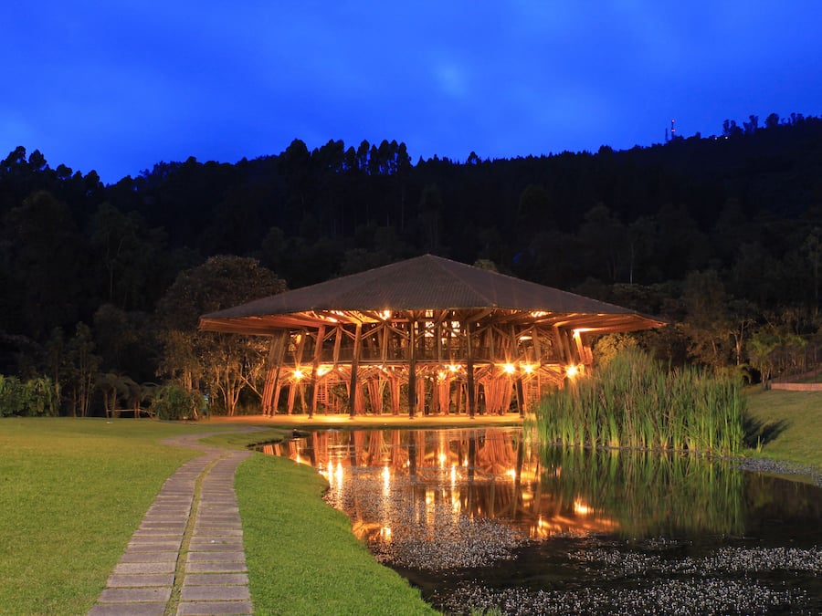 A pavilion with a roof supported by wooden beams, surrounded by a grassy area and a pond reflecting the structure.