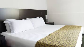 Une chambre d’hôtel avec un lit, du linge de lit blanc, un plaid à motifs, une table de chevet avec une lampe et une tête de lit sombre.