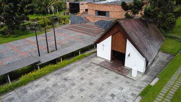 Una pequeña capilla con puerta de madera y una cruz, rodeada de un área pavimentada con un edificio de ladrillo al fondo.