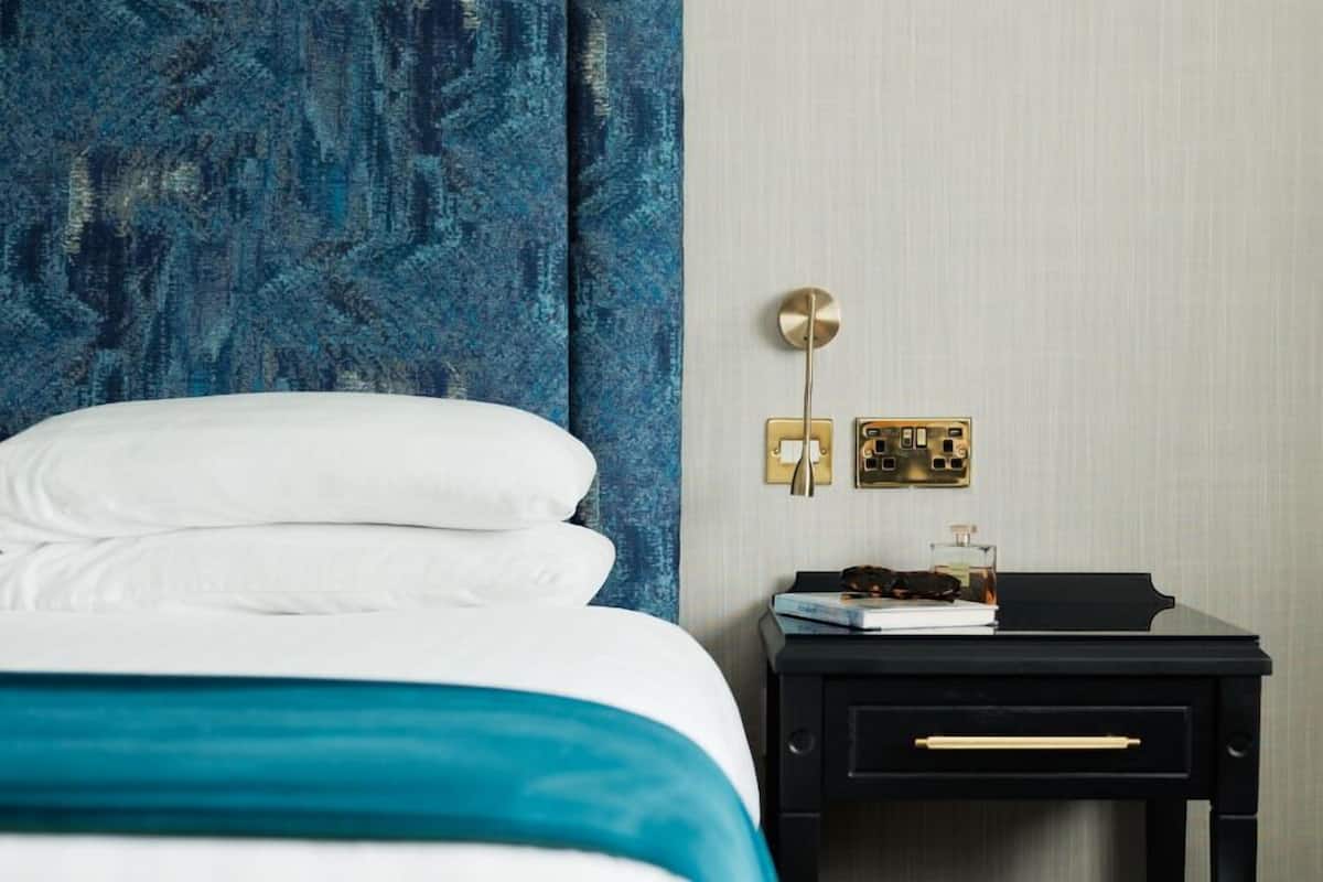 A neatly made bed with white linens, a blue accent on the bedspread, a black nightstand with a book and a bottle, and a wall-mounted light fixture.