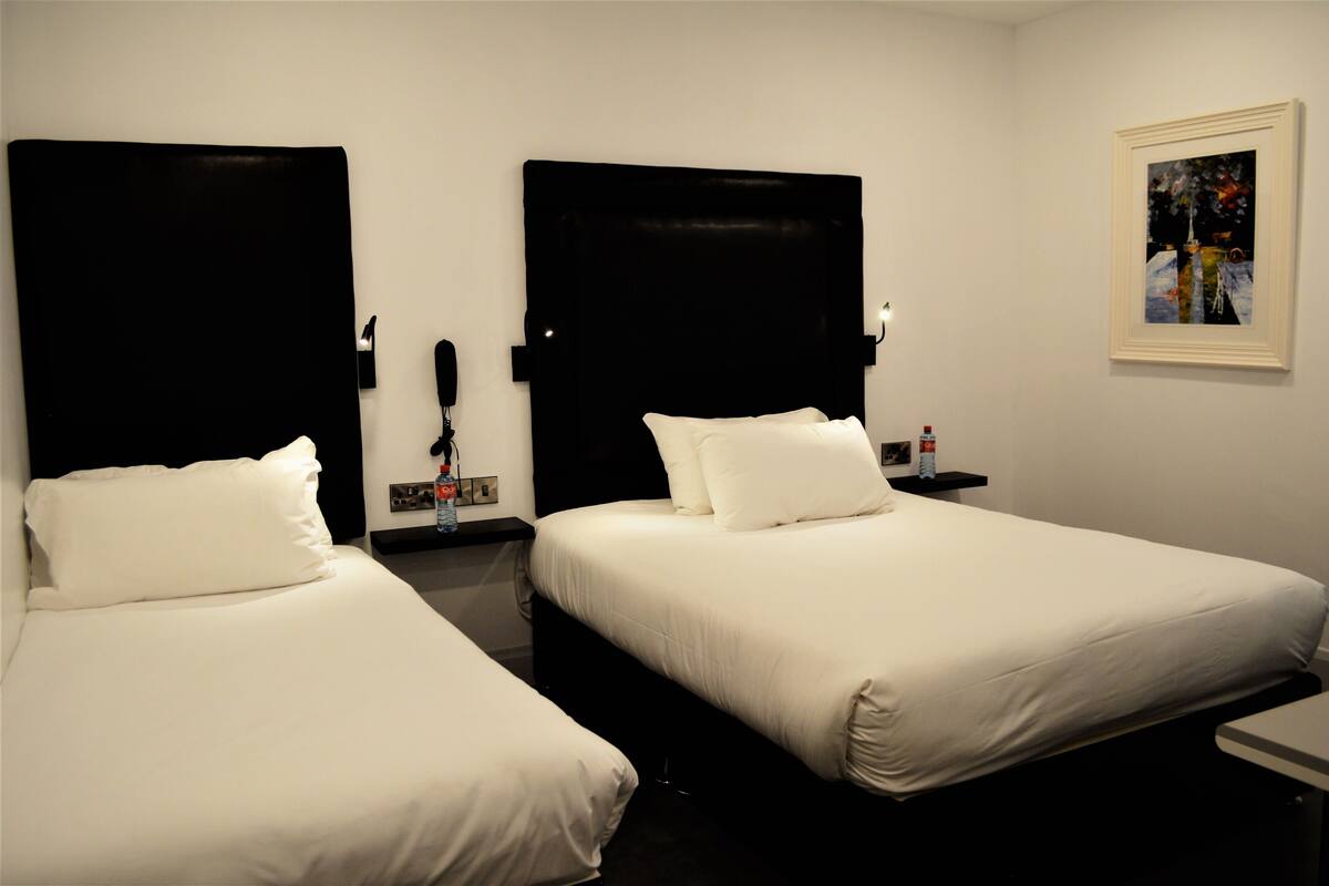 A hotel room with two beds, each with a black headboard, white bedding, and a nightstand with a phone and a bottle.
