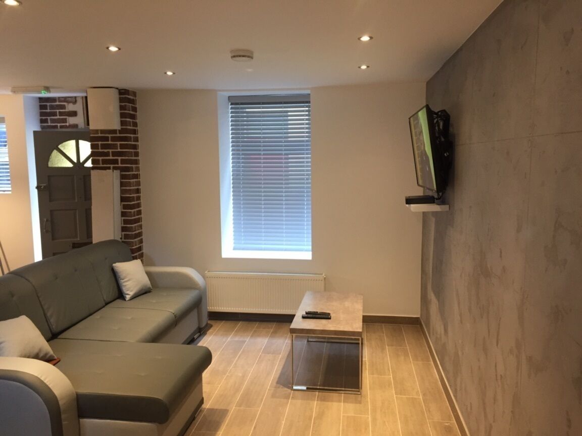 Un salon moderne avec un canapé gris, une petite table, un téléviseur fixé au mur et une fenêtre avec des stores.