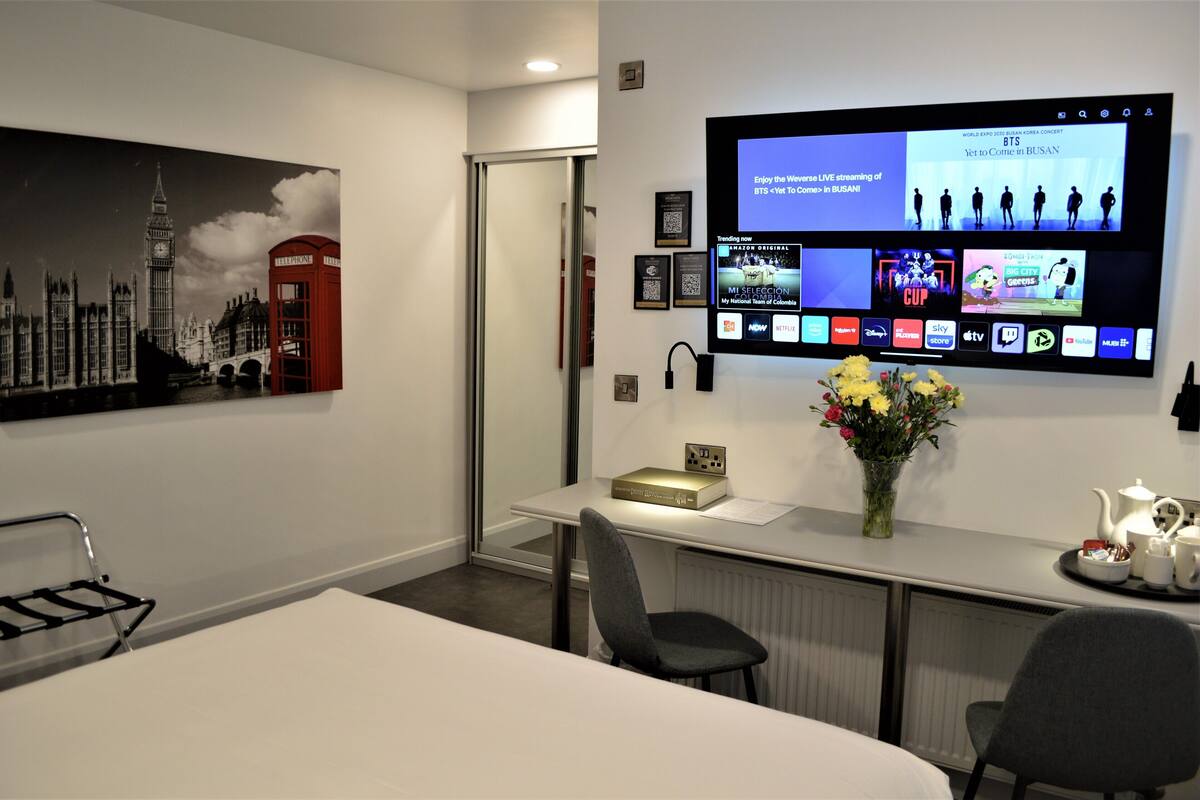 A hotel room with a large flat-screen TV displaying a live streaming service, a framed cityscape artwork, a small table with a vase of flowers, and a bed with a white comforter.