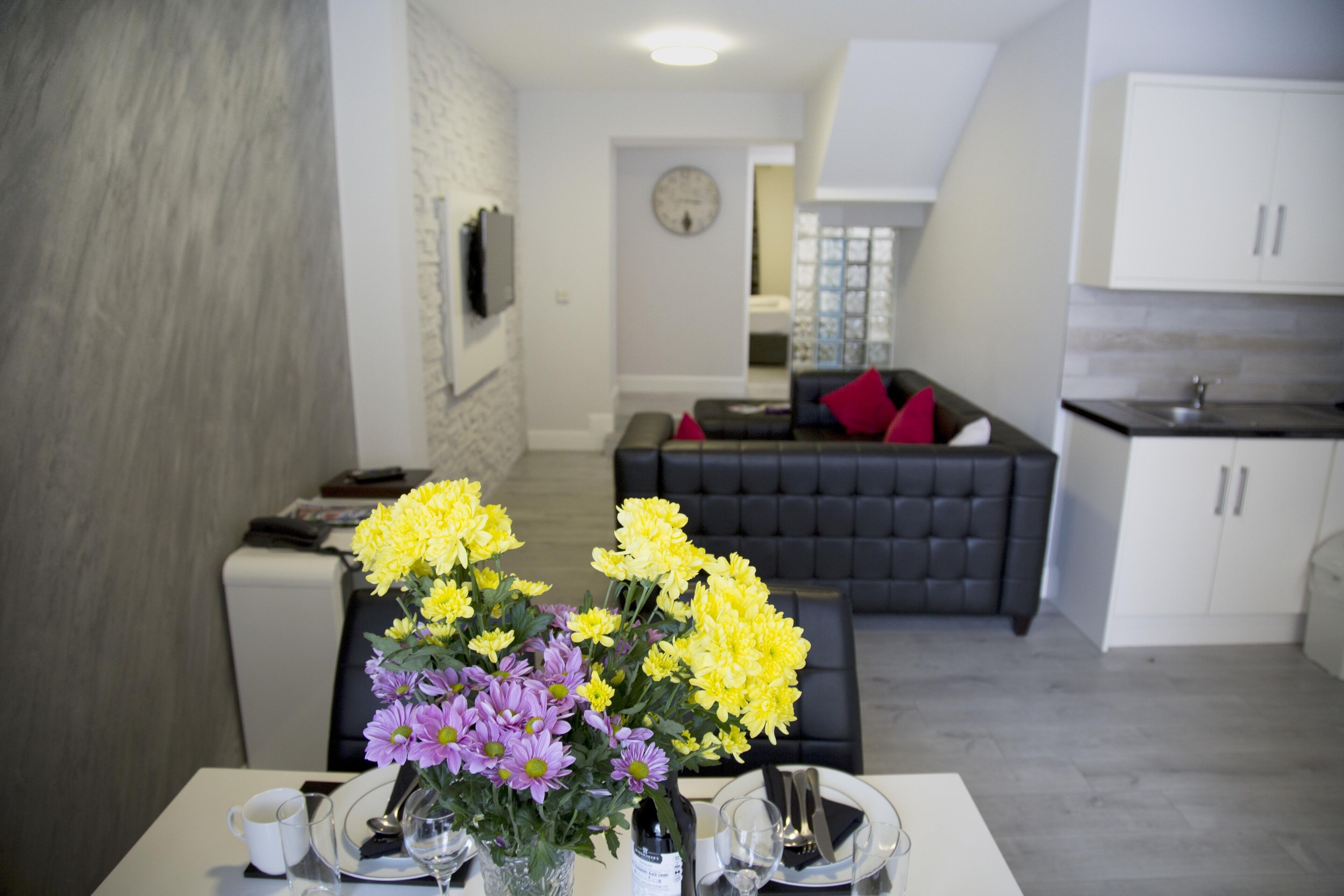 A modern living room with a dining area, featuring a sofa, a dining table set with plates, glasses, and a vase of flowers.