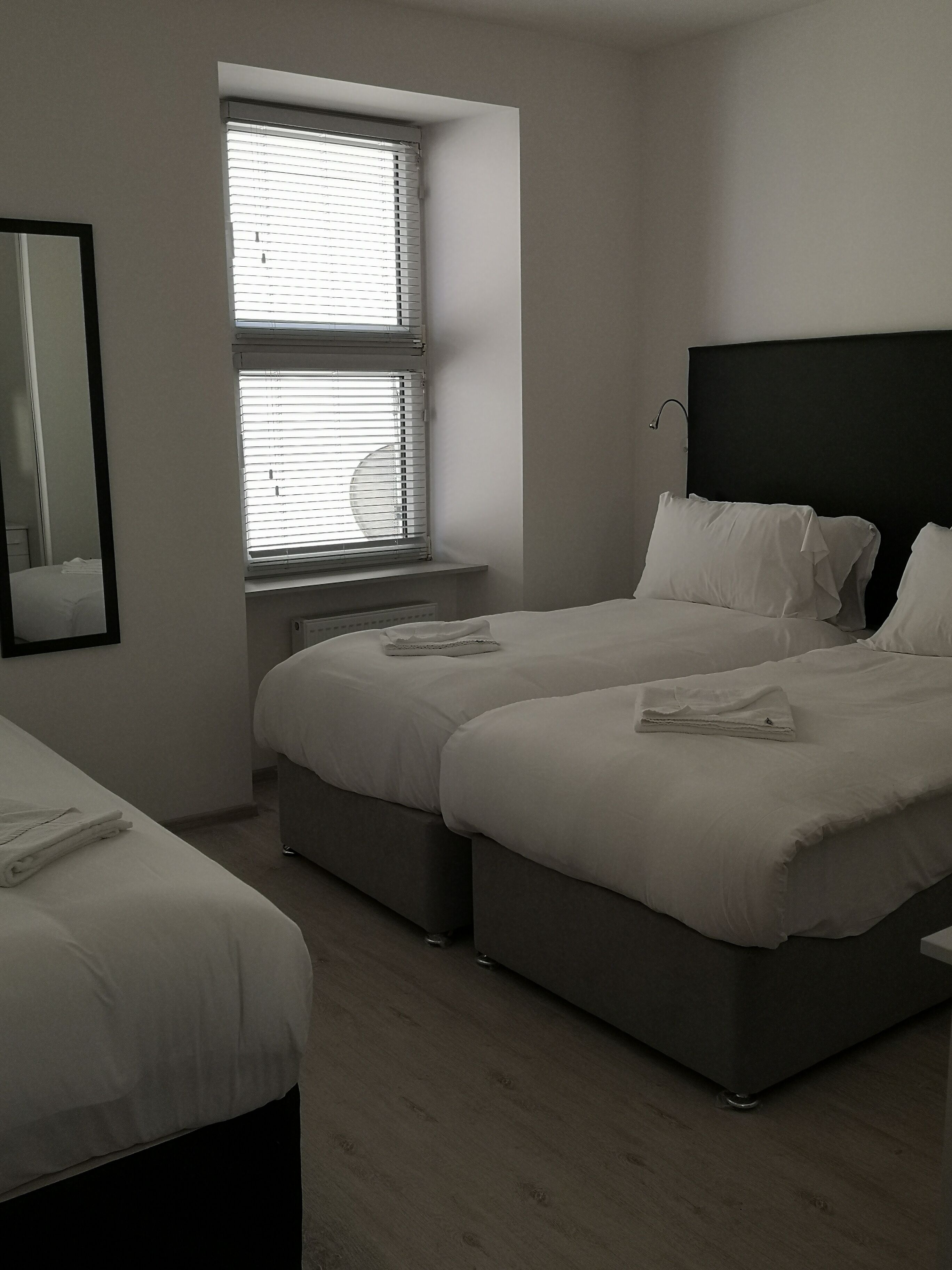 A modern hotel room with two beds, a large window with blinds, a bedside table, and a mirror on the wall.