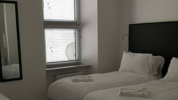 A modern hotel room with two beds, a large window with blinds, a bedside table, and a mirror on the wall.