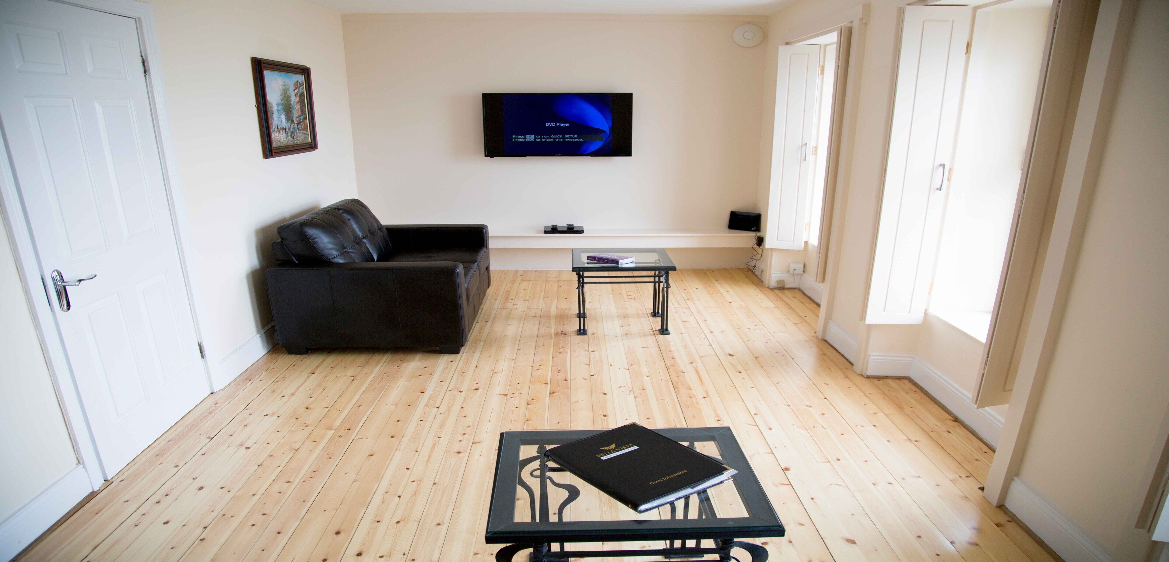 A living room with a black leather sofa, a glass coffee table, and a flat-screen TV mounted on the wall.