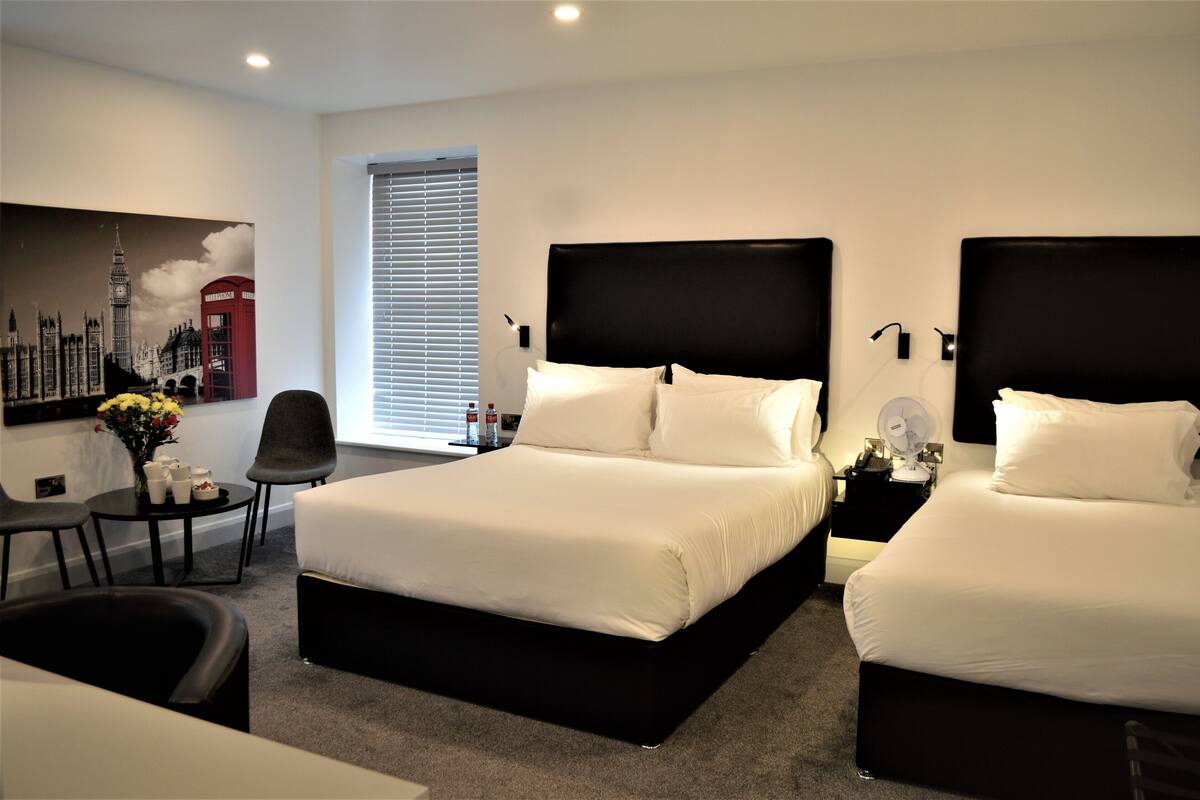 A modern hotel room with two beds, a sitting area, and a large window with blinds.