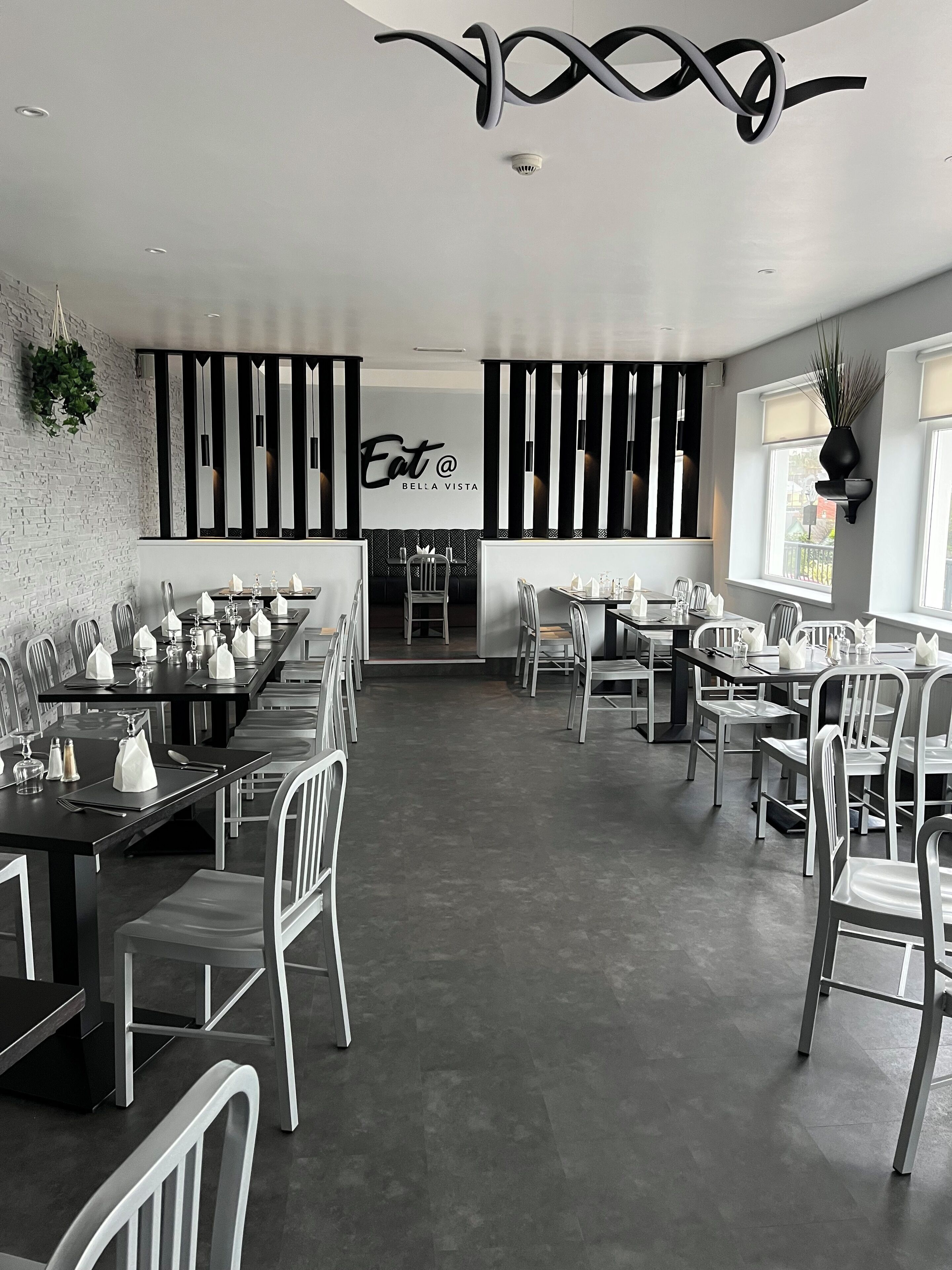 Un interior de restaurante moderno con paredes a rayas blancas y negras, un letrero central que dice 'Eat @ Bella Vista', y una larga mesa de comedor con sillas.