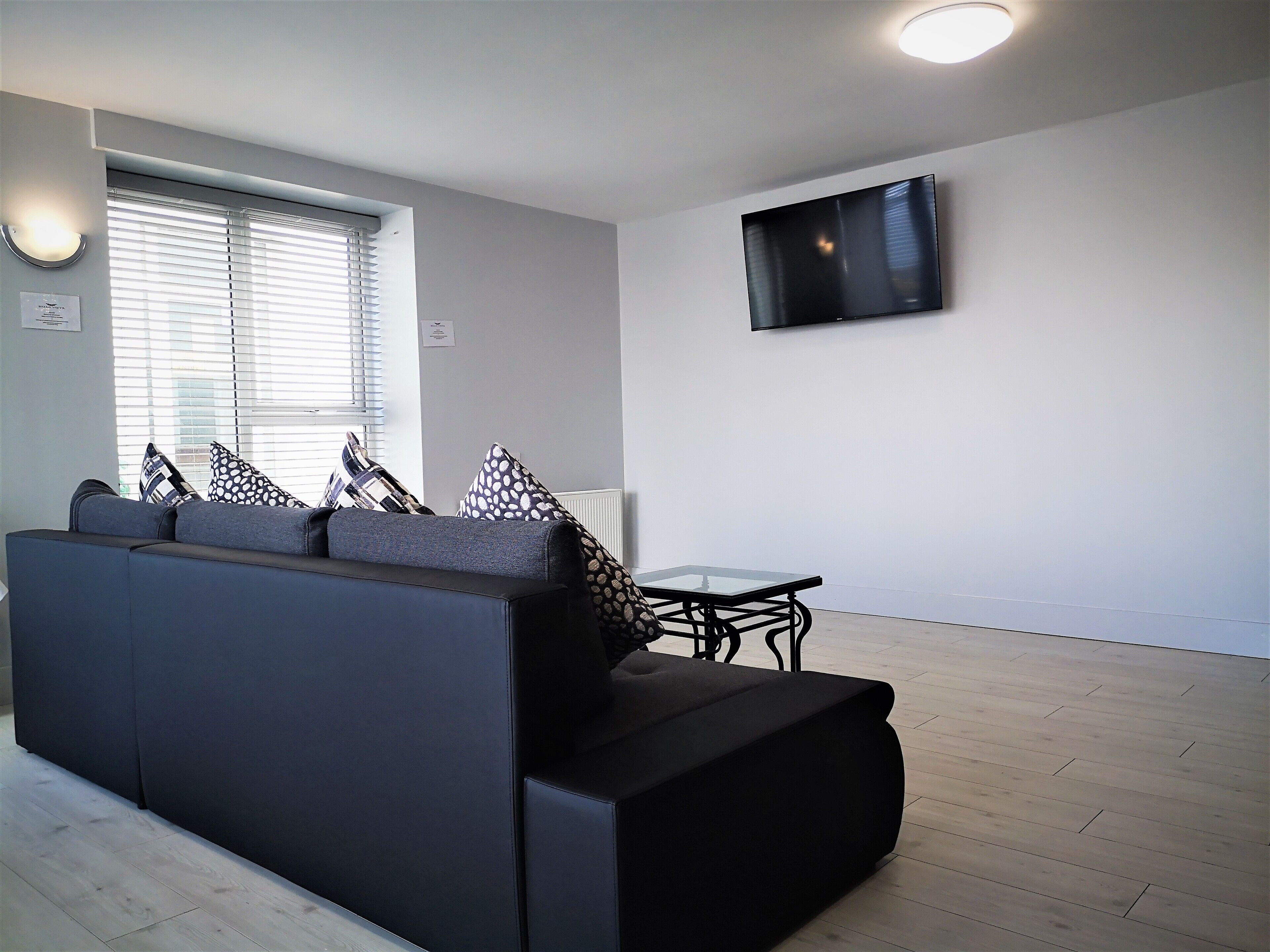 A modern living room with a dark-colored sofa, patterned cushions, a glass coffee table, and a flat-screen TV mounted on the wall.