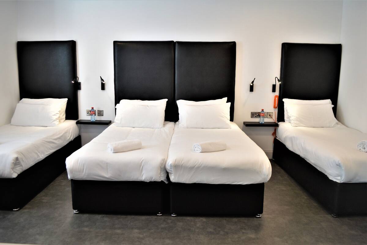 A hotel room with three single beds, each with a black headboard and white bedding. There are bedside tables with a bottle and a phone on each.