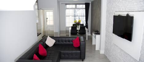 A modern living room with a black sectional sofa, a dining area with a table and chairs, and a TV mounted on a white brick wall.