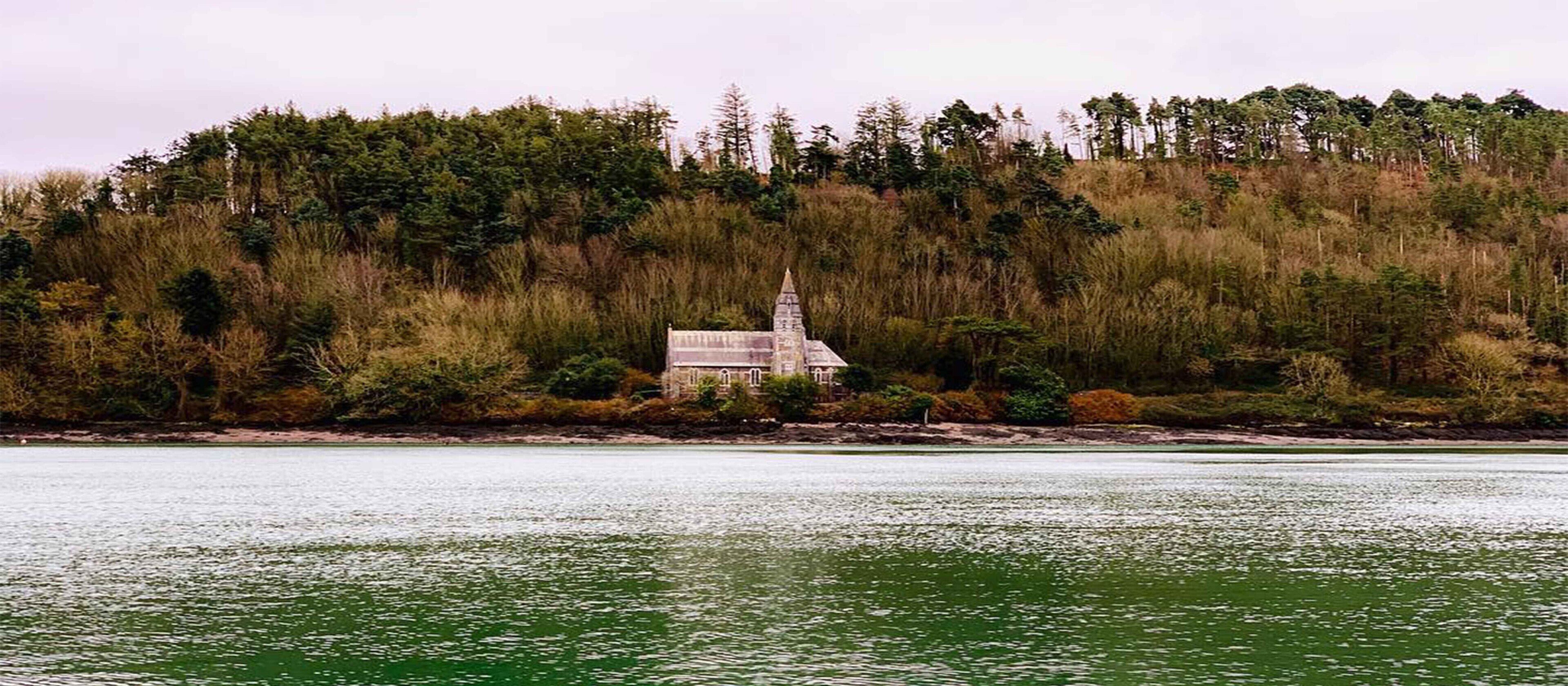 A small, isolated building with a sloped roof, surrounded by dense forest and a body of water.