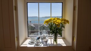 A window with a view of the sea, a vase with yellow flowers, and a tea set on the windowsill.
