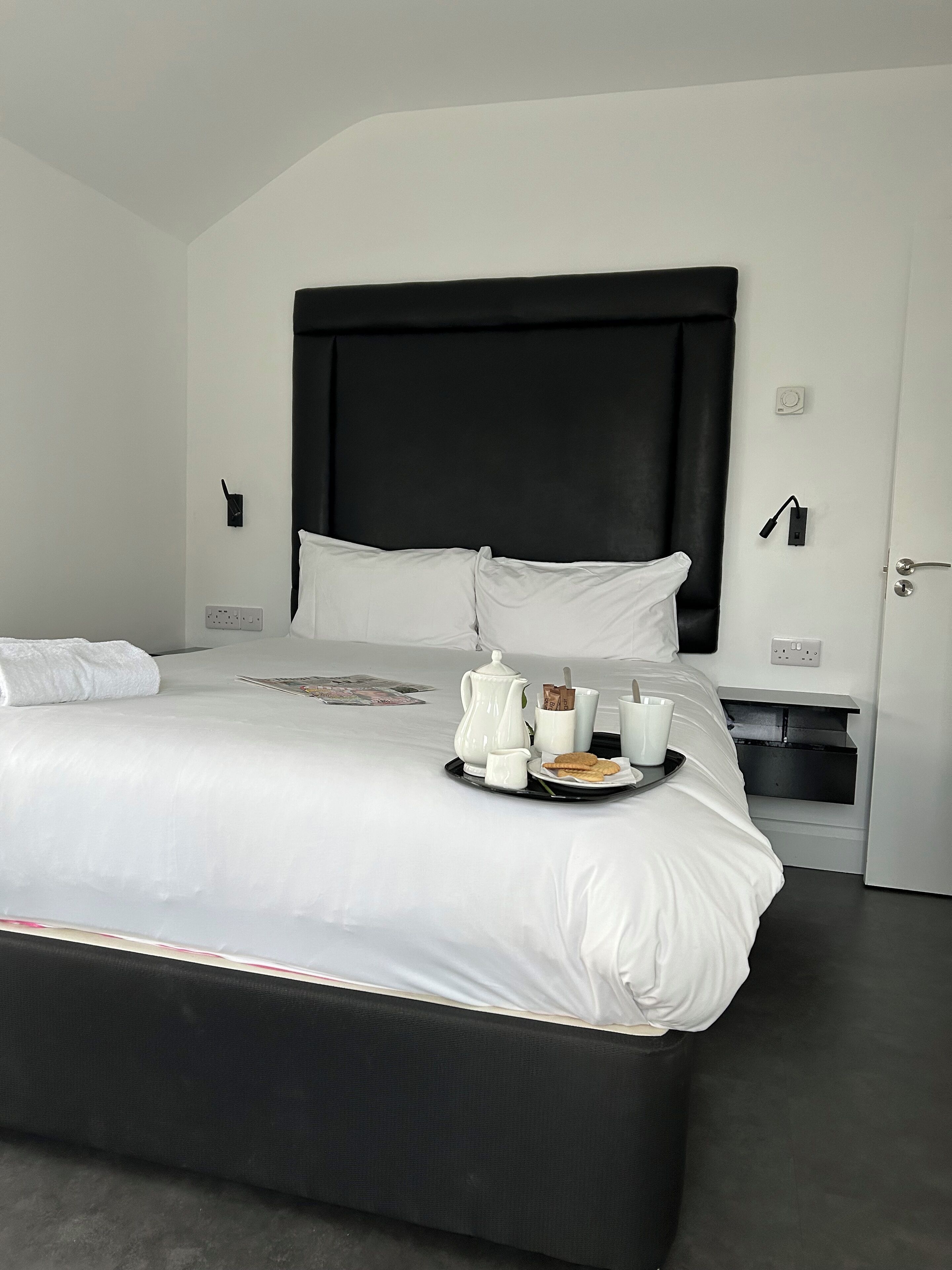 A modern hotel room with a large bed, a black headboard, white bedding, and a tray with breakfast items.