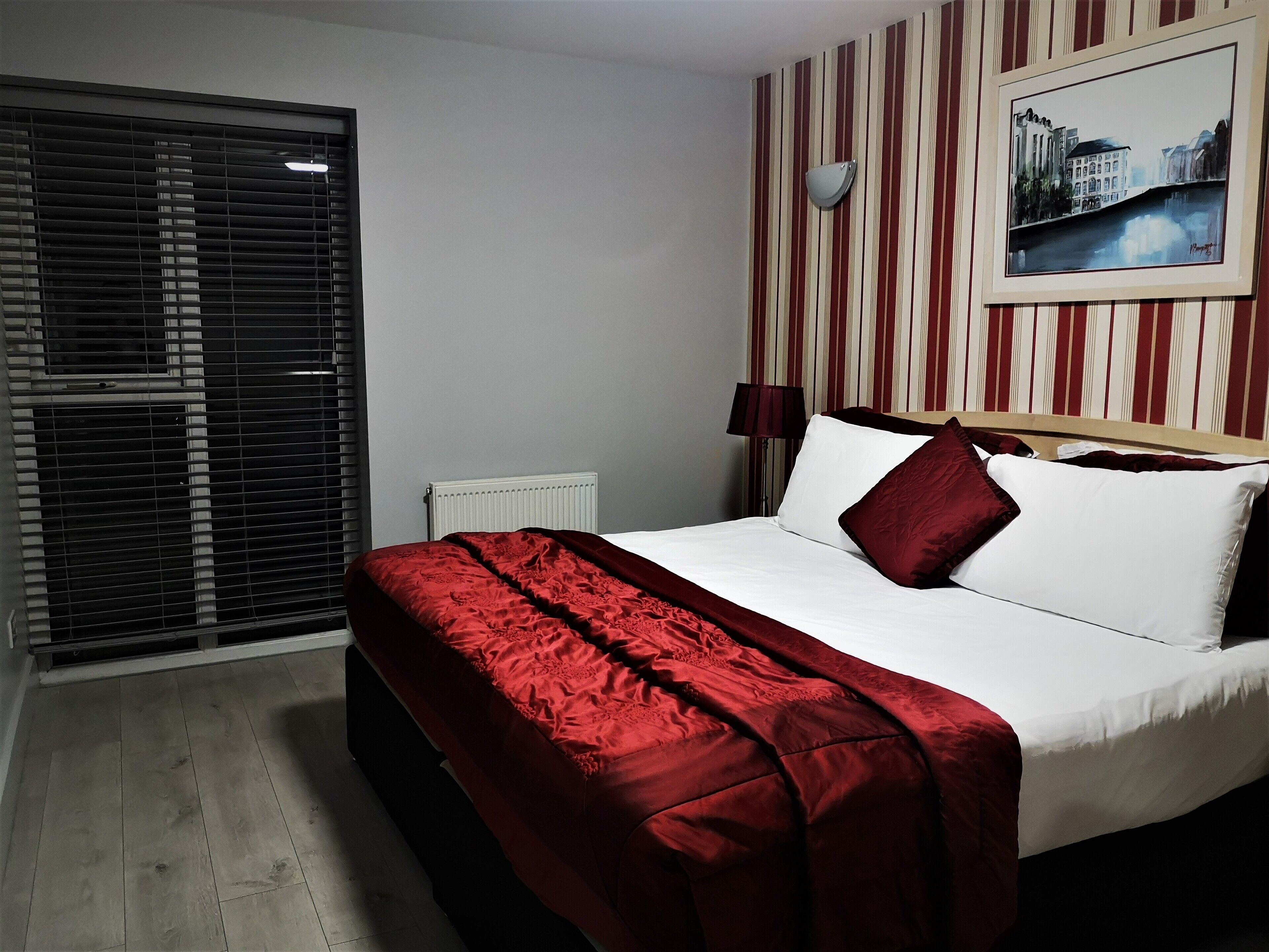 Une chambre d’hôtel avec un lit, un radiateur, une fenêtre avec des stores et un mur à rayures orné d’un tableau.