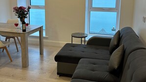 A modern living room with a grey sectional sofa, a dining table with two wine glasses and a bottle, and a vase with flowers. There are two windows providing a view of the sea.