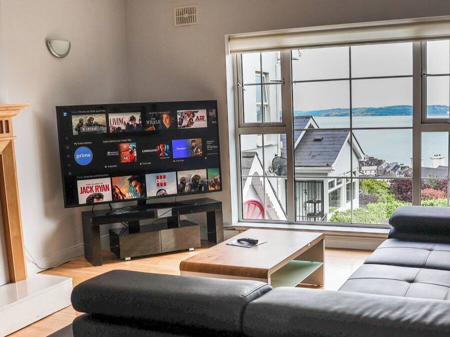 Ein modernes Wohnzimmer mit einem großen Fernseher, der verschiedene Kanäle zeigt, einem Kamin, einer Couch und Meerblick durch große Fenster.