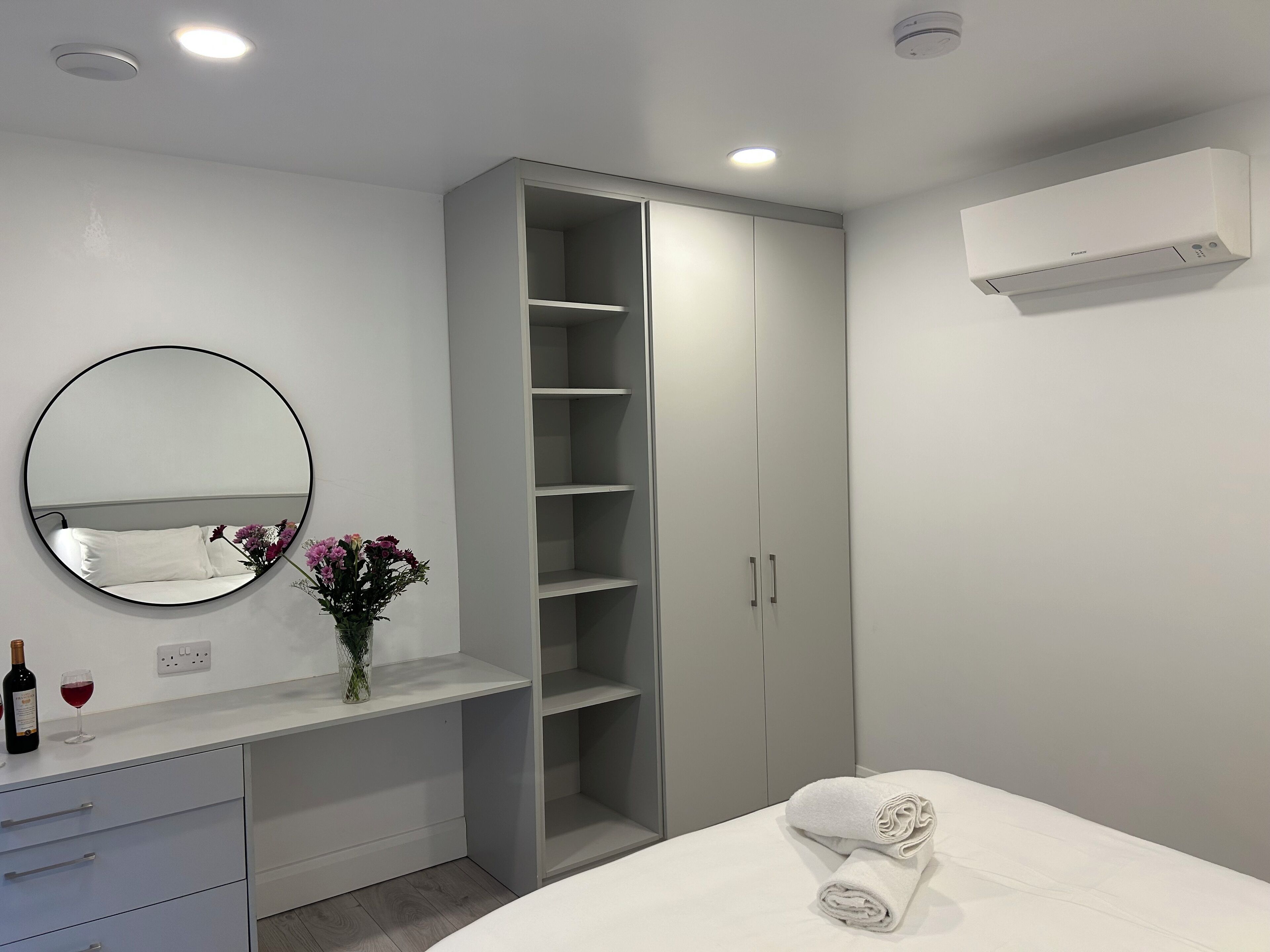 Un dormitorio con cama, armario, cómoda, espejo y un florero con flores.