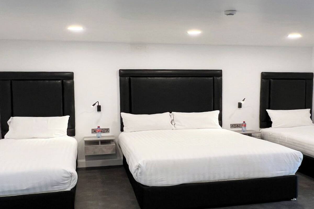 A hotel room with three single beds, each with a black headboard and white bedding. There are bedside tables with lamps and a small shelf.