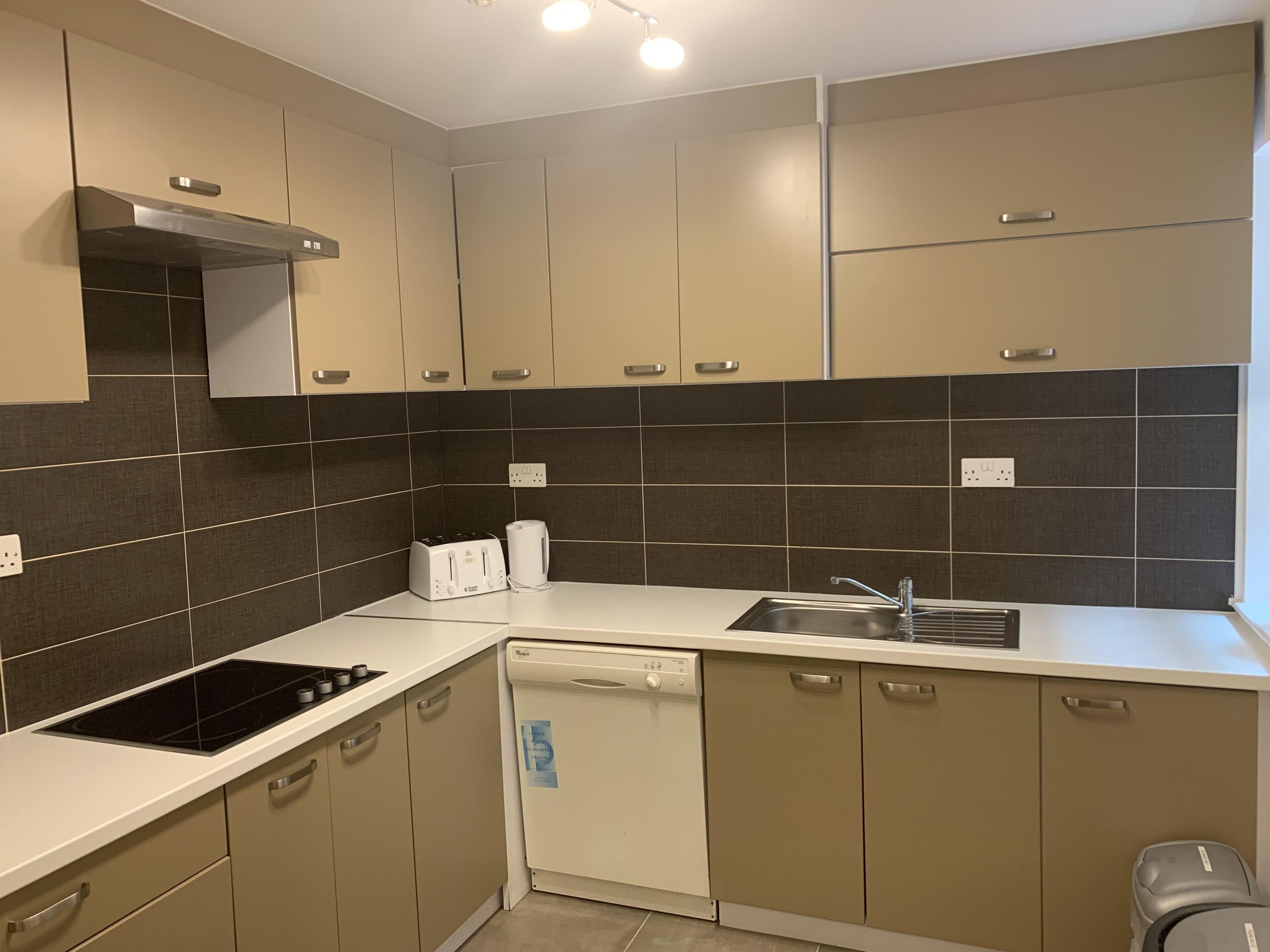 A modern kitchen with beige cabinets, a black tiled backsplash, a built-in stove, a dishwasher, and a sink.