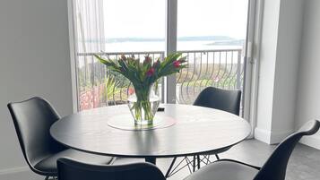 Una mesa redonda con un florero, dos sillas y una vista a un balcón con barandilla y una masa de agua al fondo.