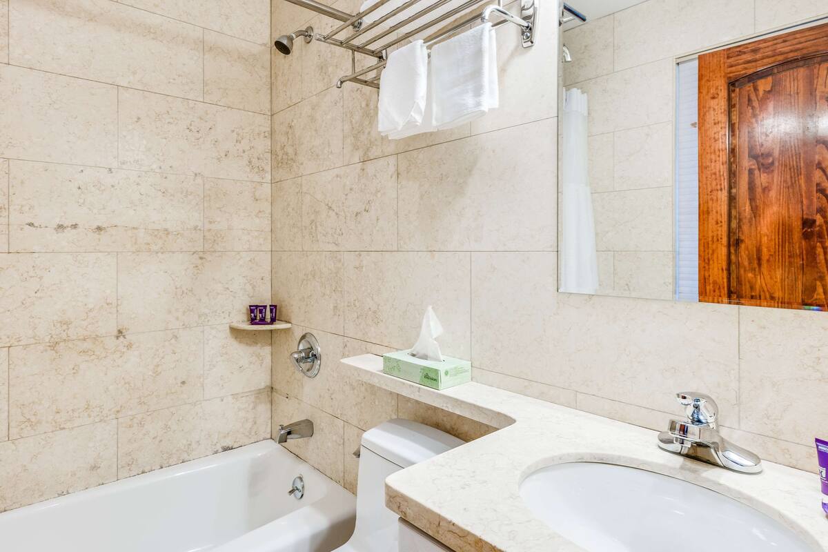 Ein Badezimmer mit einer Badewanne, einem Waschbecken und einer Toilette. Handtücher hängen an einem Halter und eine Packung Taschentücher steht auf einem Regal.