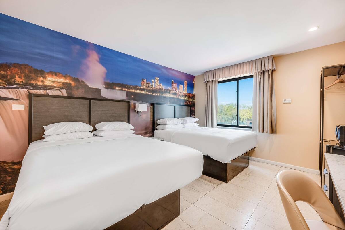 Ein Hotelzimmer mit zwei Betten, einem großen Fenster, einem Gemälde eines Wasserfalls und Blick auf eine Stadtlandschaft.