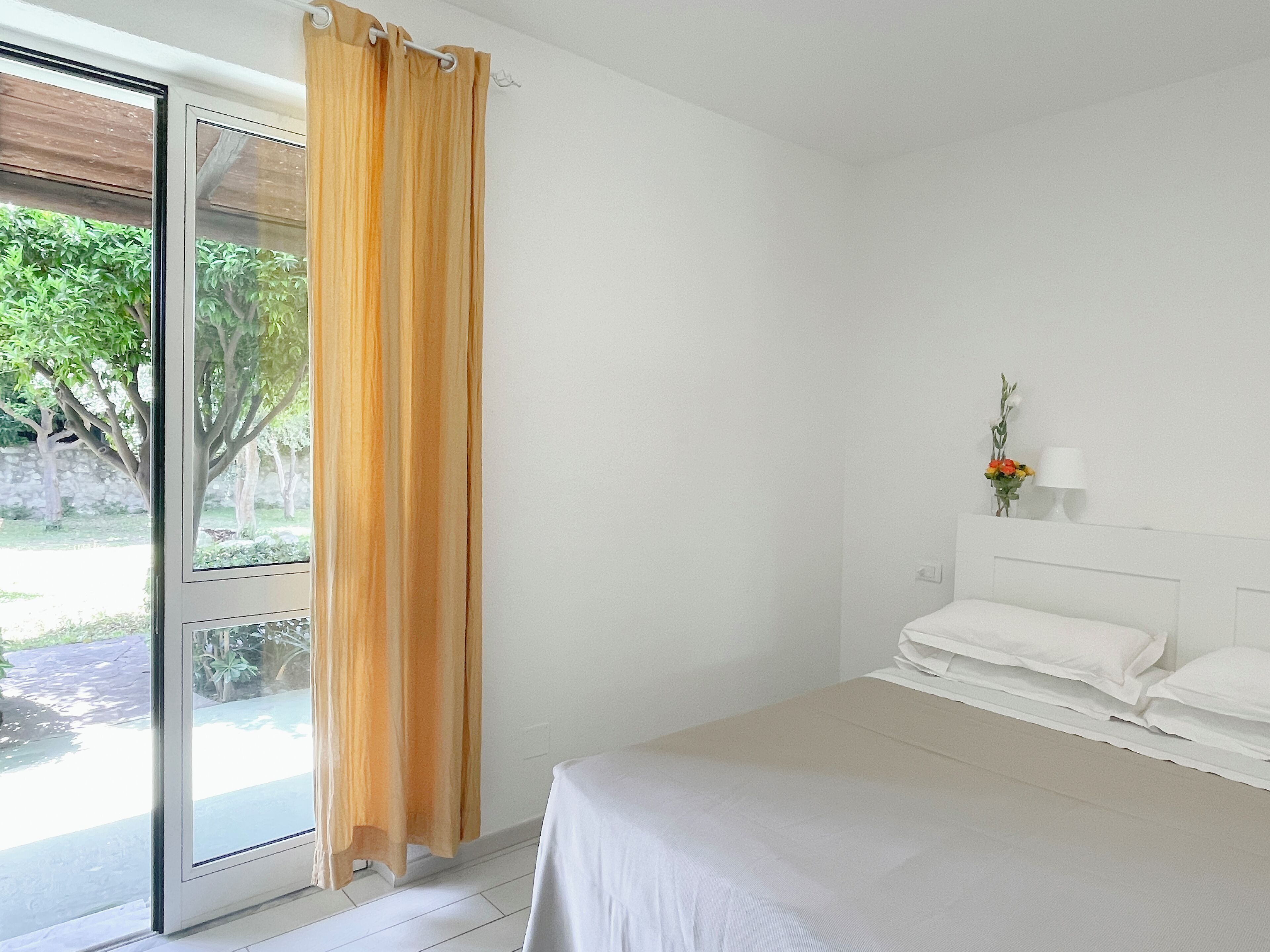 A bedroom with a bed, a window with curtains, a bedside table with a lamp, and a view of greenery outside.