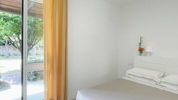 Un dormitorio con cama, ventana con cortinas, mesita de noche con lámpara y vistas a la vegetación.