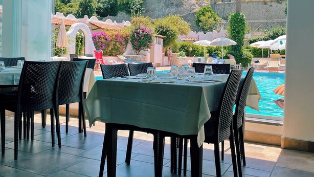 An outdoor dining area with tables and chairs, a pool, and a view of a historic tower and greenery. - Hotel Lord Byron (Forio)