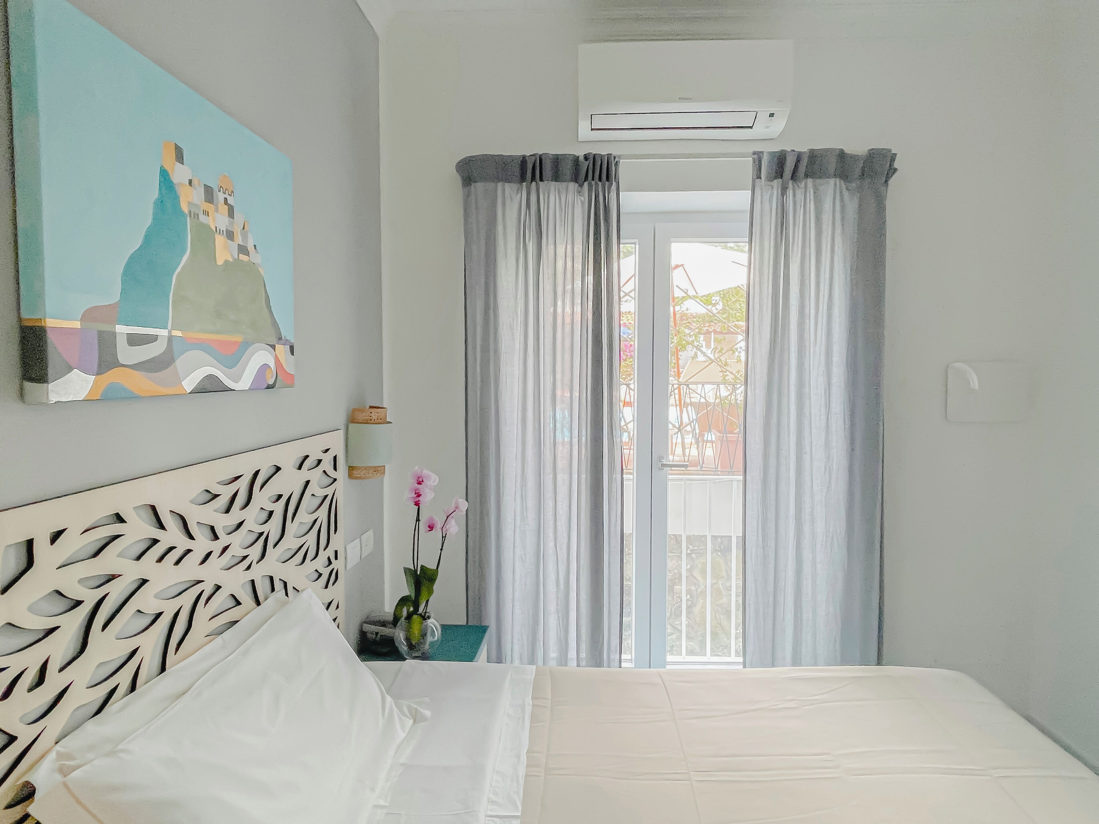 A hotel room with a bed, a painting on the wall, an air conditioning unit, and a view through the window.