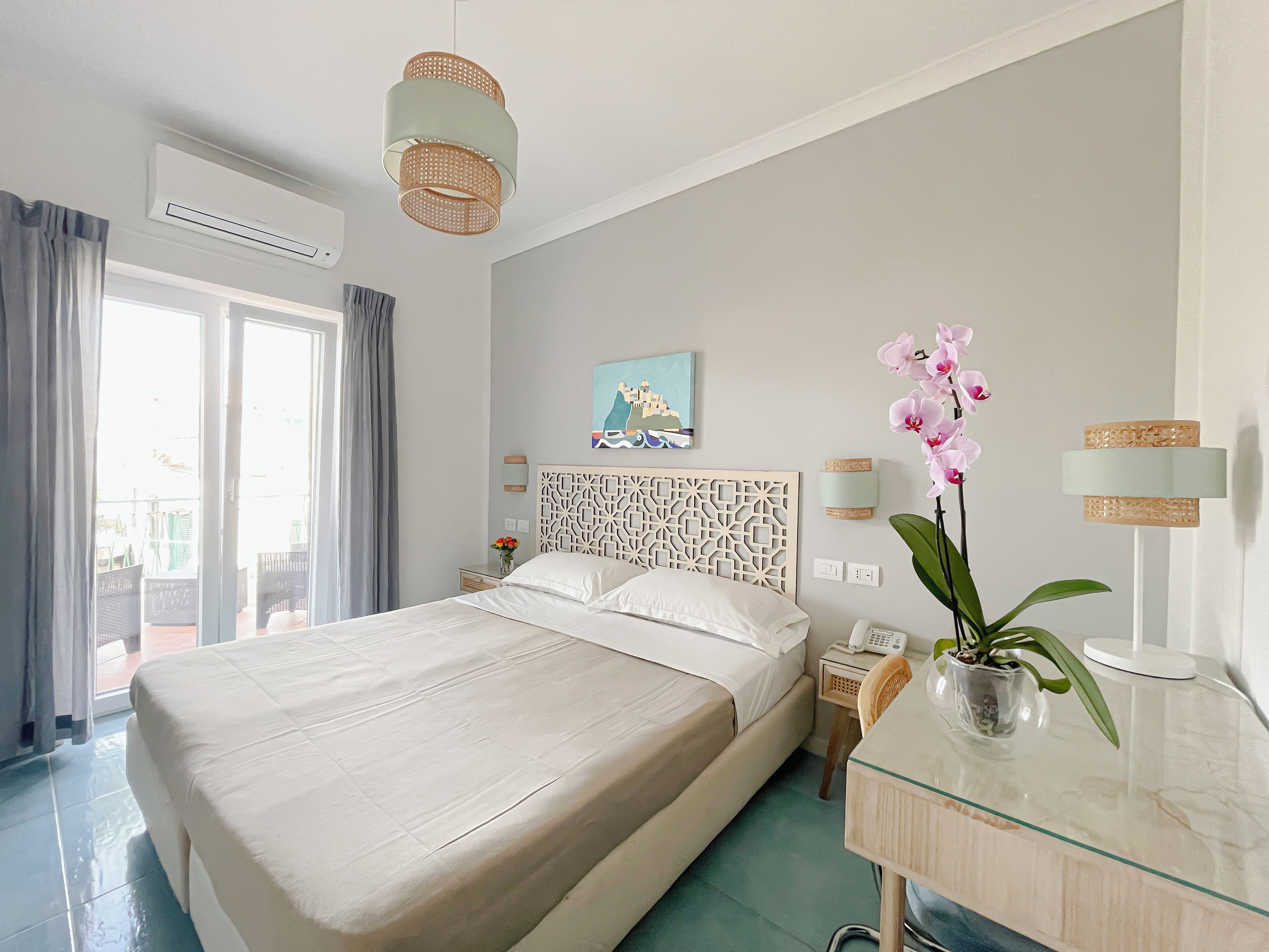 A hotel room with a large bed, a bedside table with a phone, a vase with flowers, and a painting on the wall.