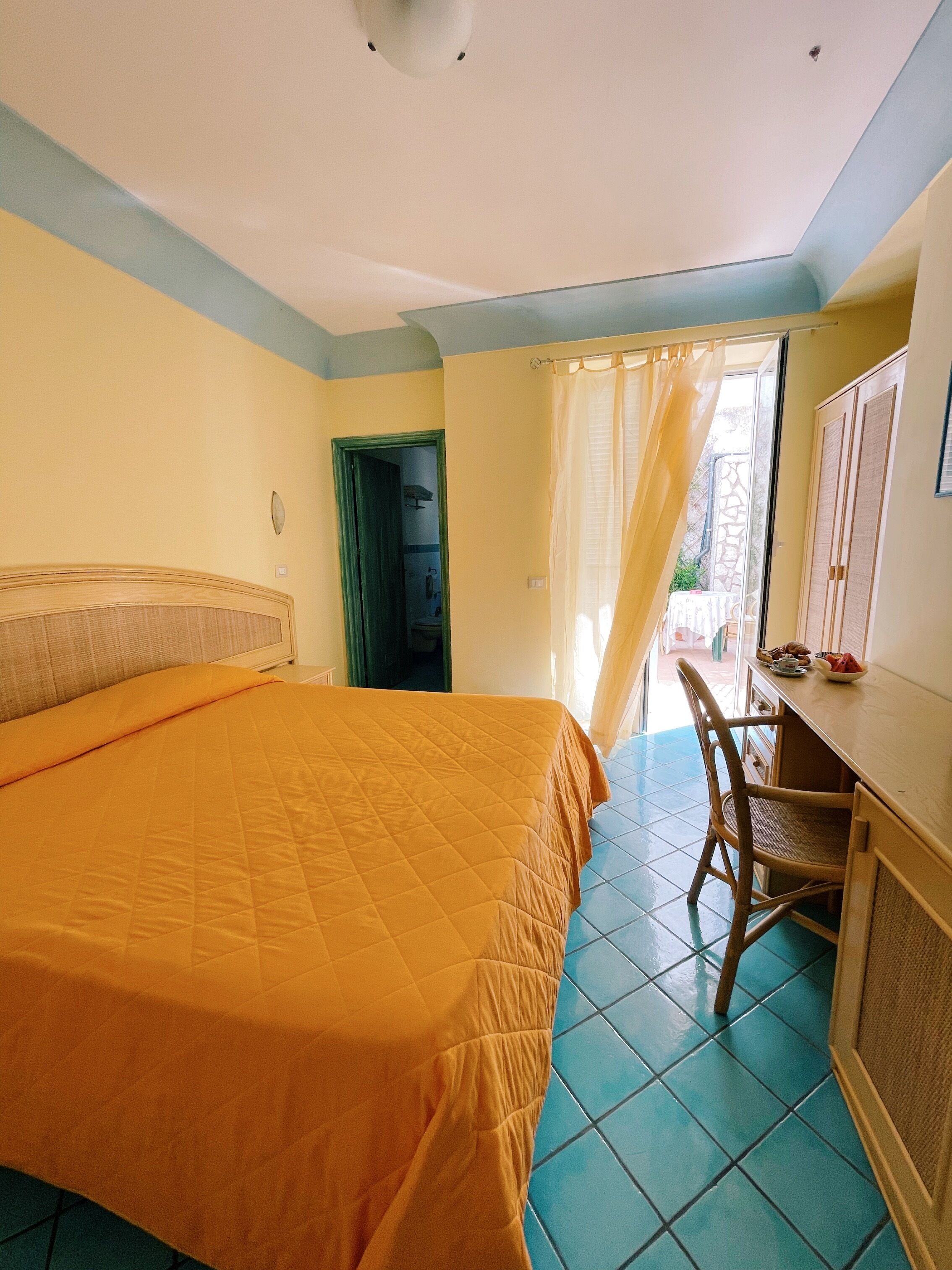 Une chambre avec un grand lit, un bureau et une chaise. Il y a une porte qui donne sur une autre pièce et une fenêtre avec des rideaux.