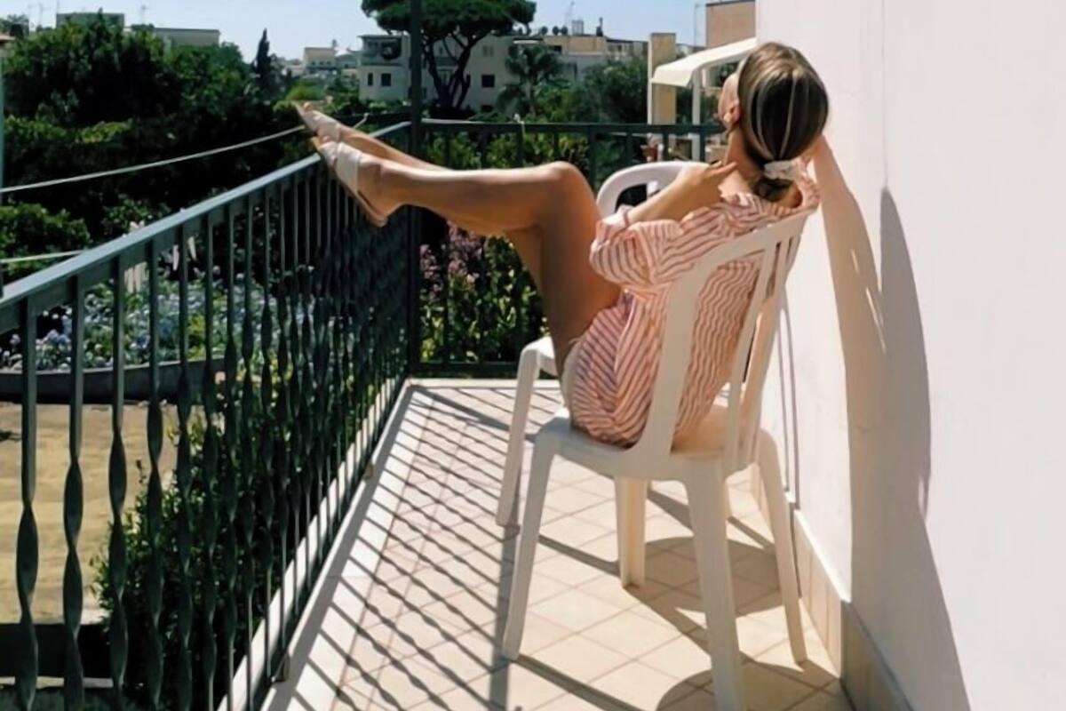 Une personne assise sur une chaise, sur un balcon, d’où l’on peut admirer des bâtiments et des arbres.