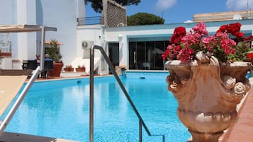A swimming pool with a stone planter with flowers, a metal ladder, and a building with a balcony in the background.