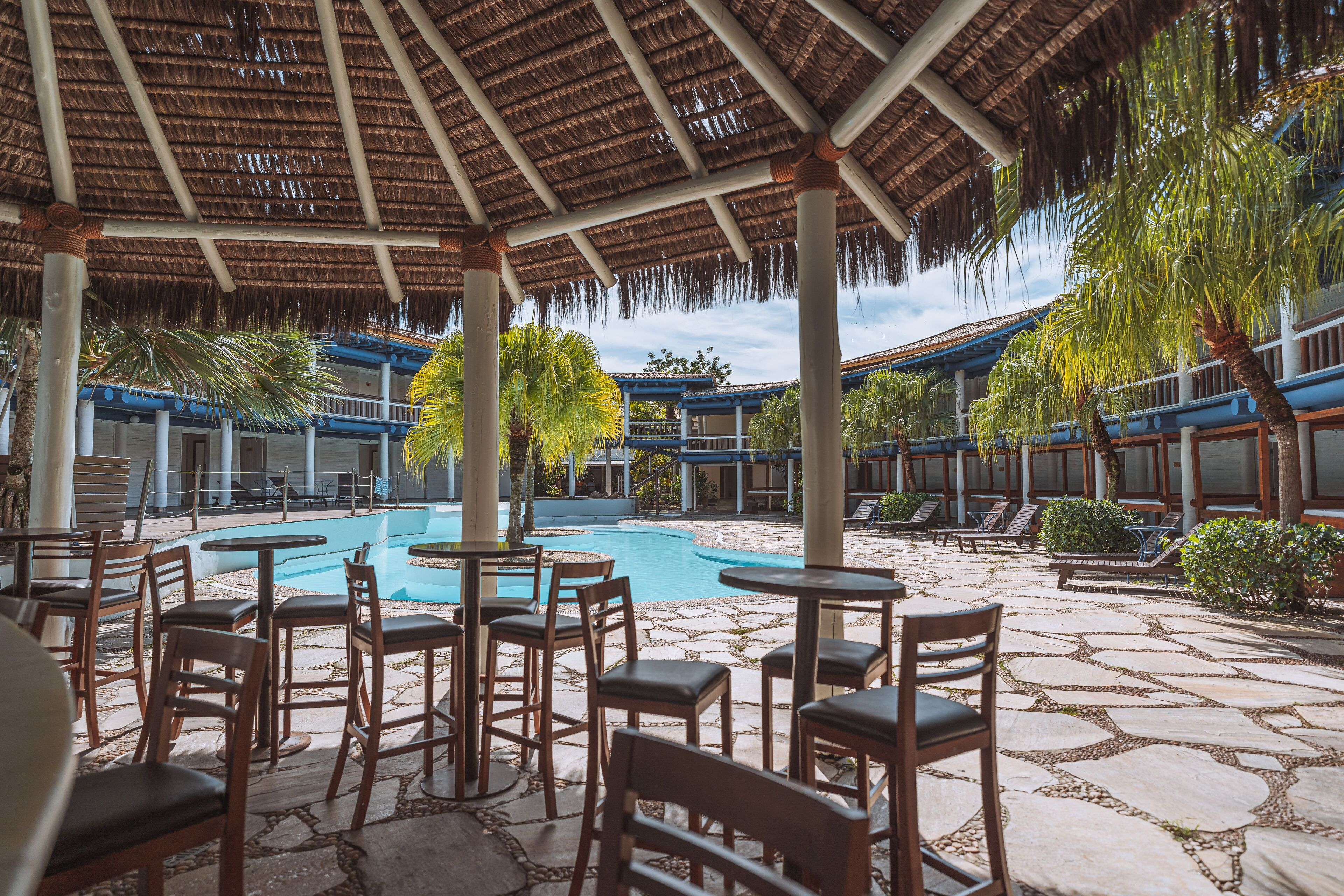 A pool area with lounge chairs and tables, surrounded by stone flooring and palm trees.