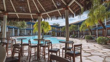 A pool area with lounge chairs and tables, surrounded by stone flooring and palm trees.