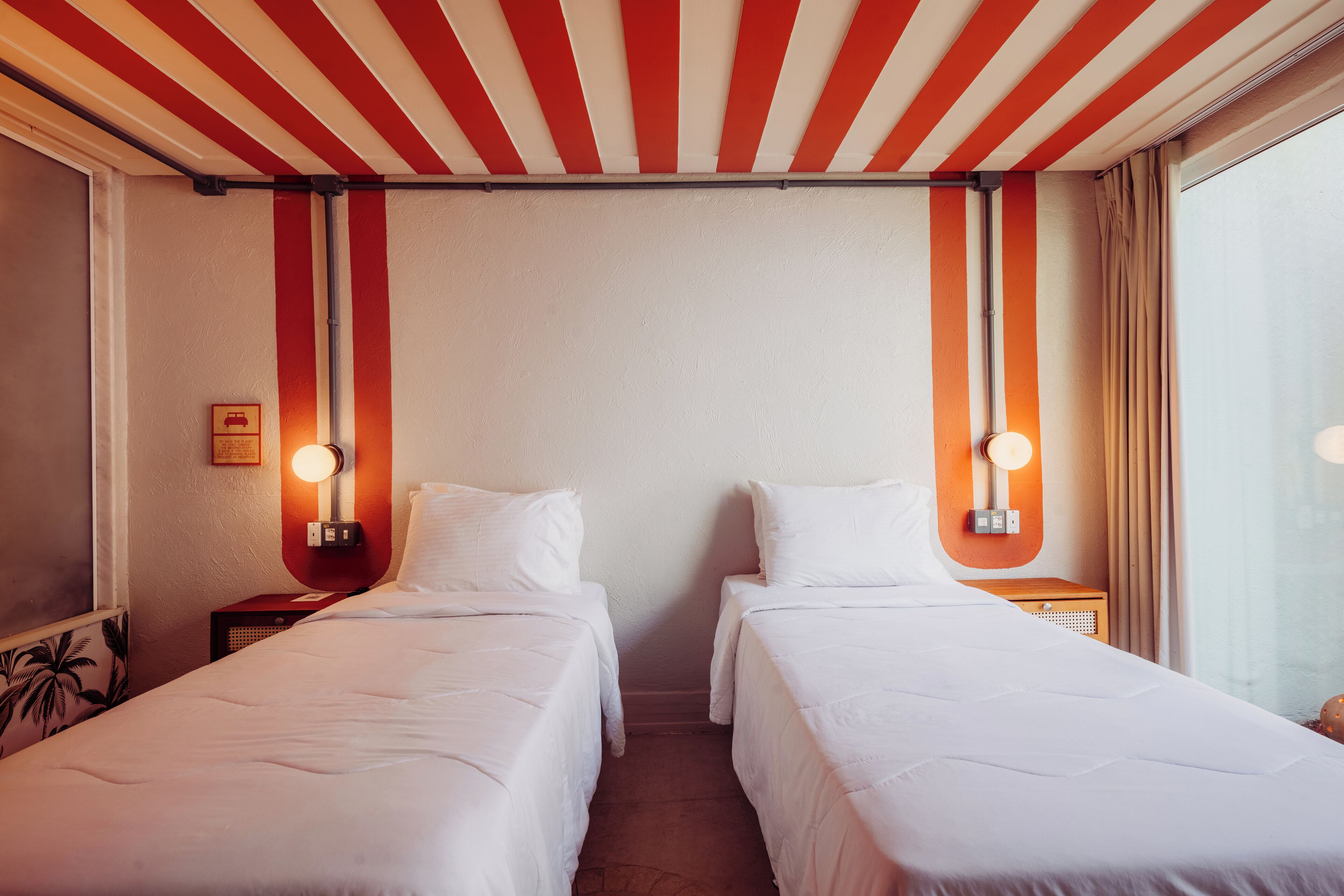 Habitación de hotel con dos camas individuales, techo a rayas rojas y blancas, y lámparas fijadas a la pared.