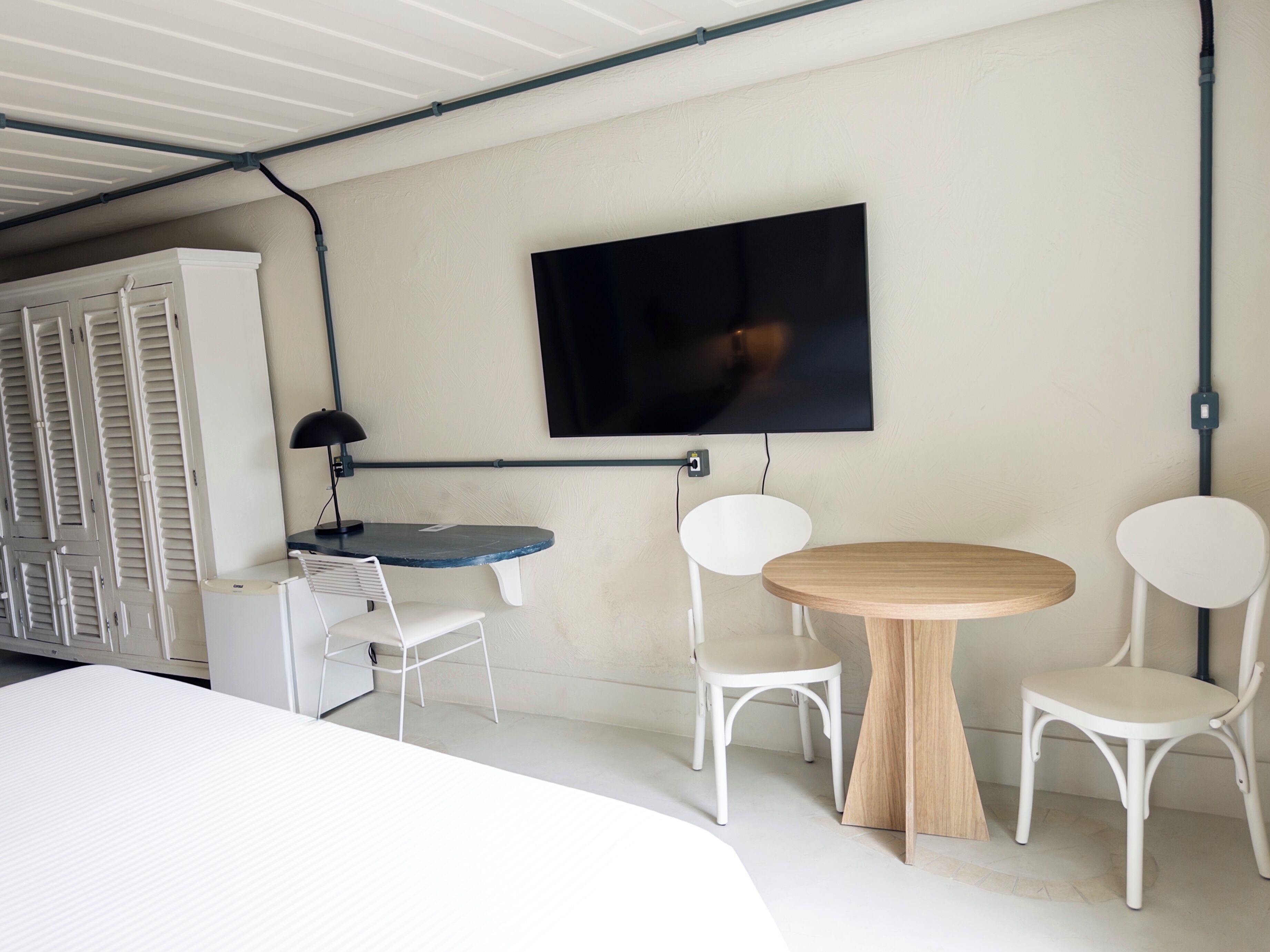A modern room with a bed, a desk with a chair, a round wooden table with two chairs, a TV mounted on the wall, and a wardrobe with louvered doors.
