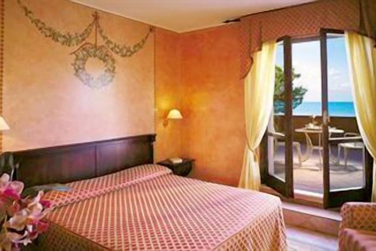 A hotel room with a bed, a nightstand, a lamp, and a view of the ocean through a glass door.