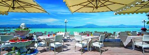 An outdoor dining area with tables and chairs, a large planter with plants, and a view of a body of water and mountains in the background. - Hotel Lugana Parco Al Lago (Sirmione)