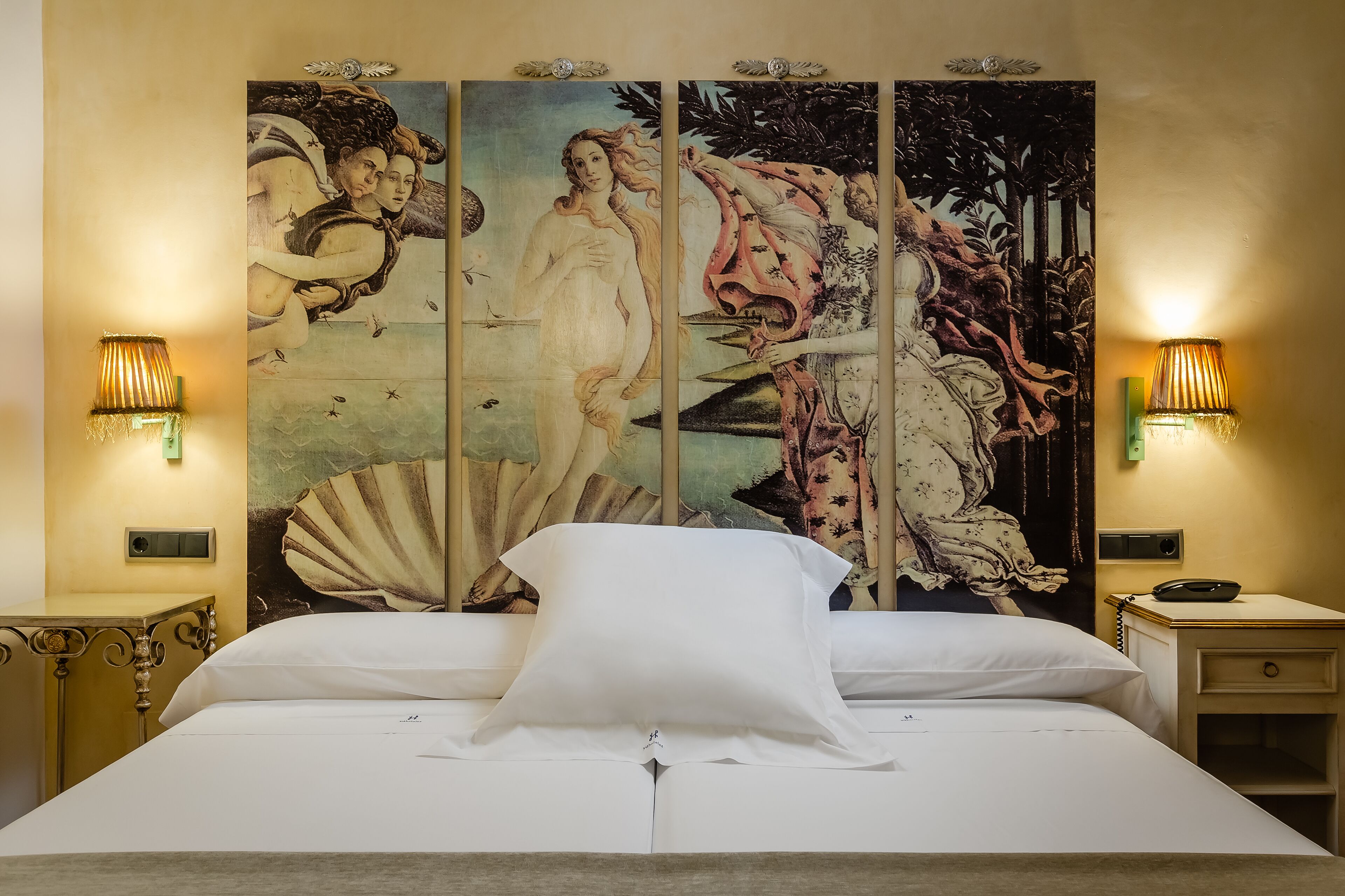 A bed with white linens, flanked by two nightstands with lamps, and a large, ornate tapestry on the wall behind.